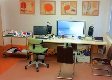 Modernes Arztpraxis-Büro mit Computer, Schreibtisch, Stühlen und medizinischem Wagen.